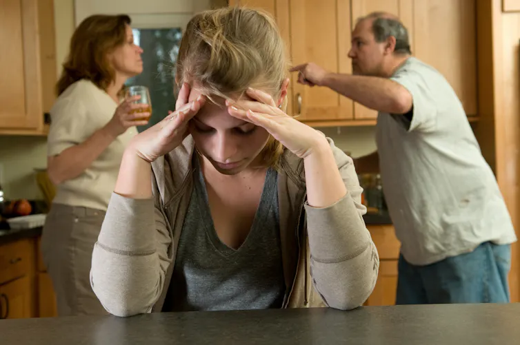 a young girl sits with her head in her hands as two adults yell ateach other in the background