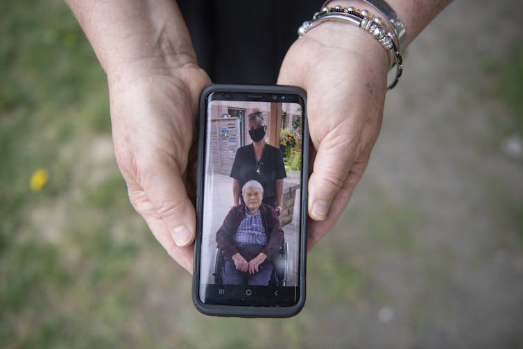 two hands holding a phone. The screen shows an elderly man in a wheelchair and a woman wearing a face mask standing behind him