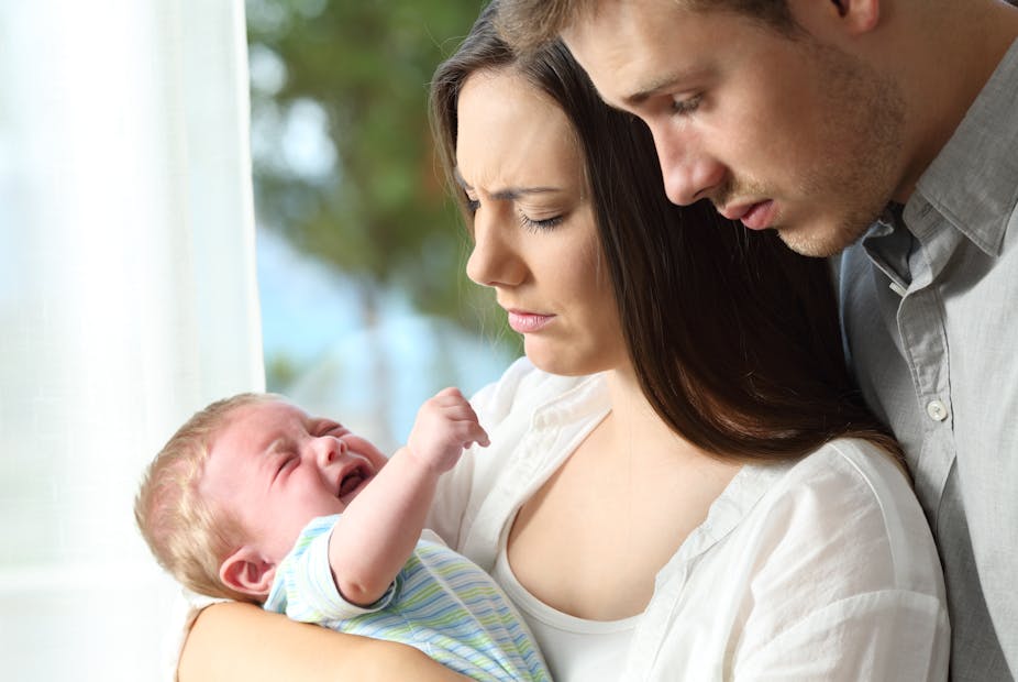 A tired and sad couple look at their crying baby, held by the mother.