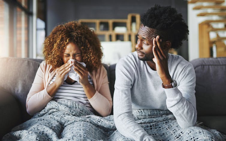 A woman and a man sit on a couch under a blanket. The woman is sneezing.
