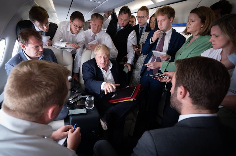 Boris Johnson sitting on an aeroplane surrounded by reporters.