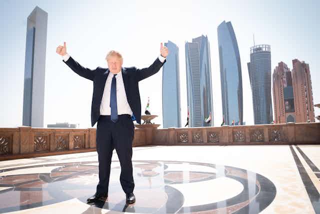Boris Johnson give a thumbs up but has a sad face while posing on top of a building in front of skyscrapers in Abu Dhabi