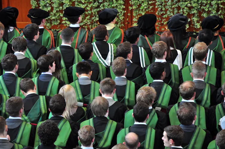 Students in gowns stand in rows for a university graduation ceremony.