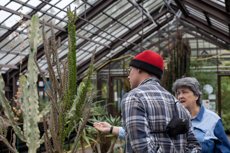 A woman gestures at succulent plants in a greenhouse while a man looks on.