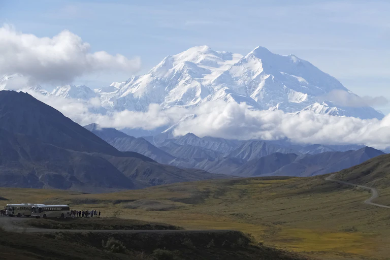 A snow-capped mountain in the distance behind green tundra and a tour bus.