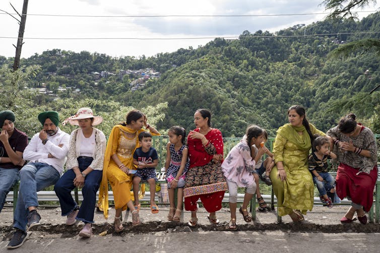 About half a dozen people in brightly colored clothes sit and chat on a fence with hills in the background.