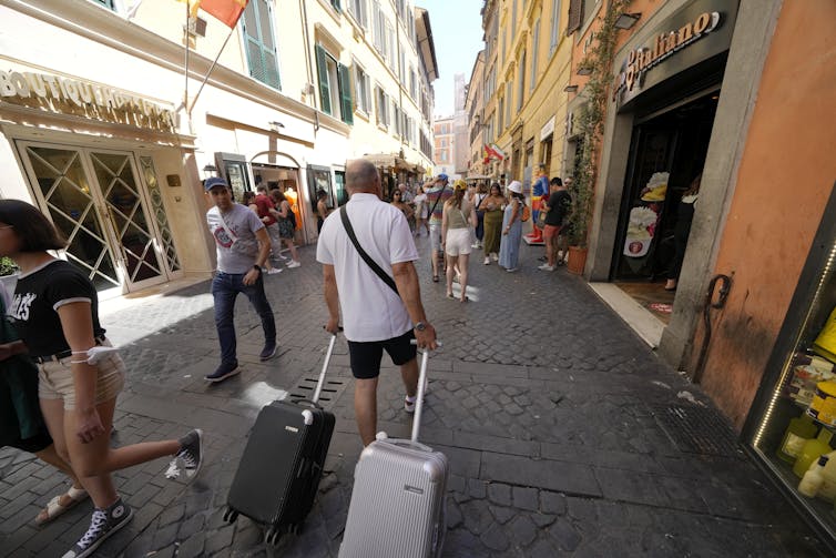 A man in a white shirt drags two small suitcases along a small, crowded sidewalk.