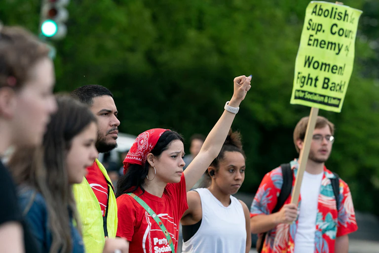 A row of young people appear to protest, with one woman wearing a red bandana raising her fist. A man at the end of the line has a yellow sign that says, 'Abolish Supreme Court Enemy, women fight back.'