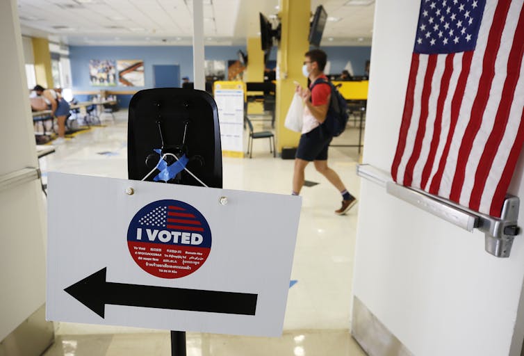 A sign says 'I voted' in a voting room, with one young man walking holding a ballot