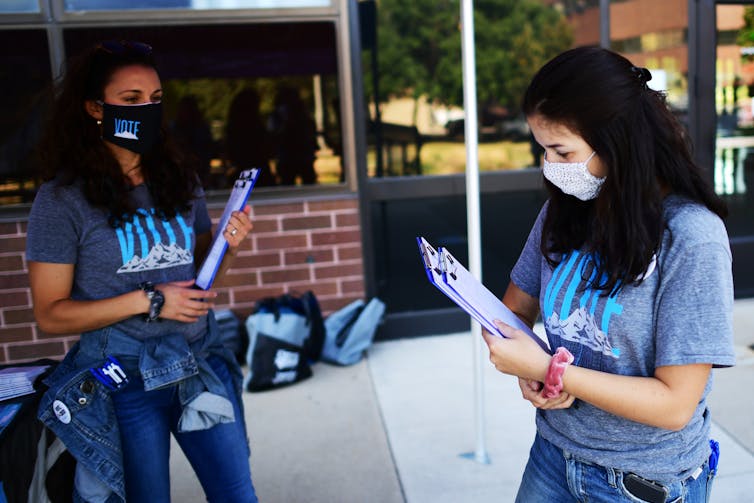 Two young women wear blue shirts and face masks and hold clipboards.