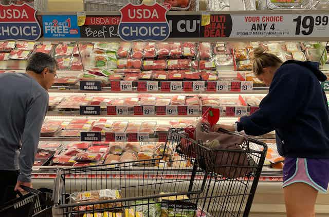 Two shoppers peer into a meat refrigerator with sales prices above it.