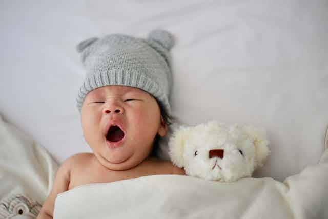 A baby in a grey hat yawns with a white grumpy teddy bear.