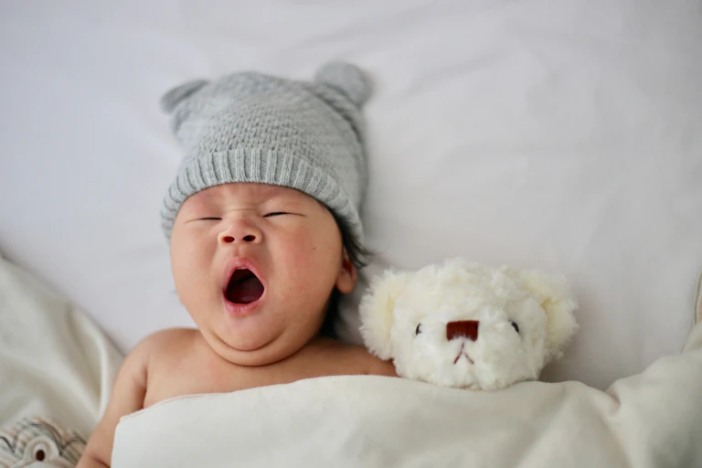 A baby in a grey hat yawns with a white grumpy teddy bear.