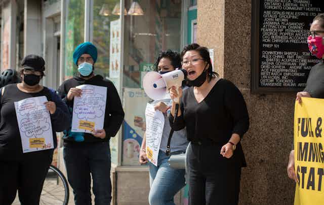 A woman speaks into a megaphone as people stand around her carrying signs. One man wears a teal turban.