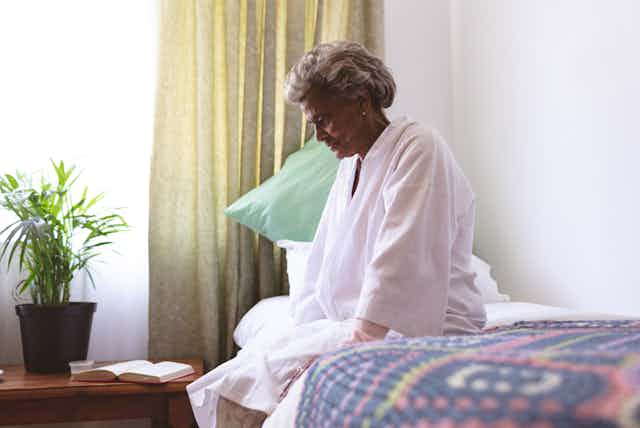 Older woman wearing a white robe sitting on a bed looking pensive