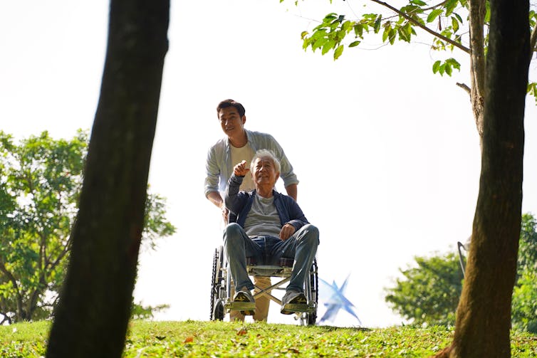 A young man pushing an older man's wheelchair between two trees in a grassy area