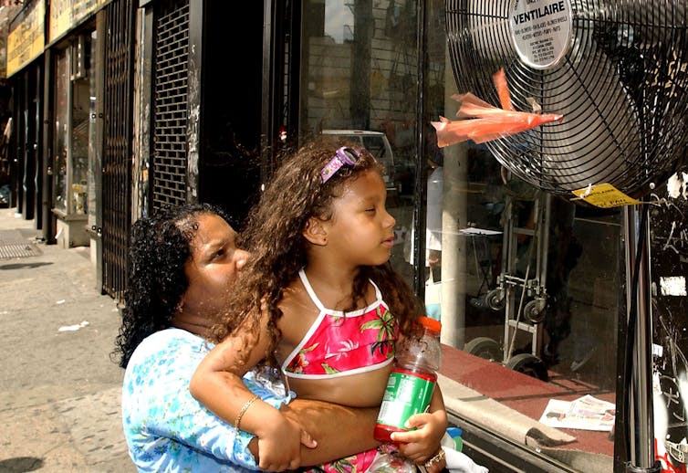 A woman holds a young girl up to a fan in front of a store on a hot day.