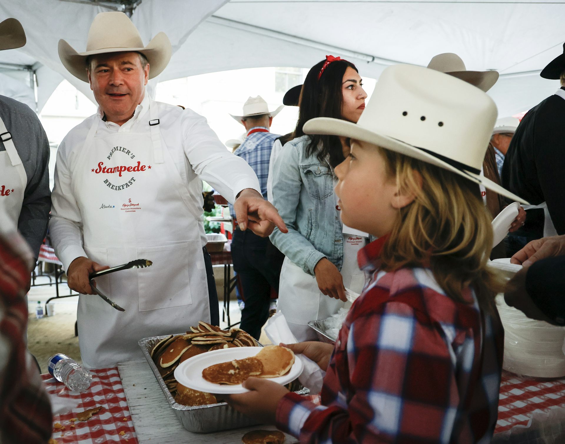 A man in a white cowboy hat and apron that says Premier's Stampede Breakfast serves pancakes to a child. The child also wears a white cowboy hat.