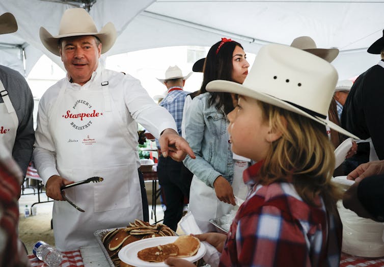 A man in a white cowboy hat and apron that says Premier's Stampede Breakfast serves pancakes to a child. The child also wears a white cowboy hat.