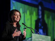 A dark-haired woman smiles as she stands behind a microphone.
