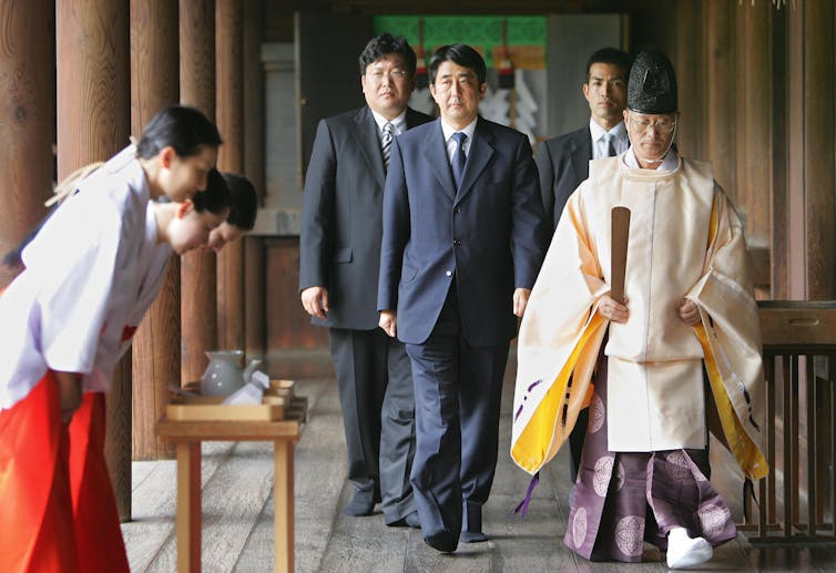 Three men in black suits walk through a hallway with a priest ahead of them and three others bowing to them.