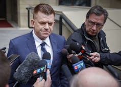 A man in a blue suit stands surrounded by CTV microphones.