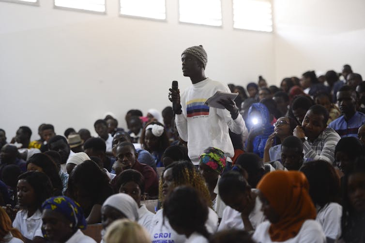 A black student in a white t-shirt and a knit cap speaks into a microphone among other students.