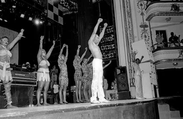 Black and white photo of a theatre stage. A shirtless man stands in front of a row of women in African fabric stage costumes, all raising both hands in fists.