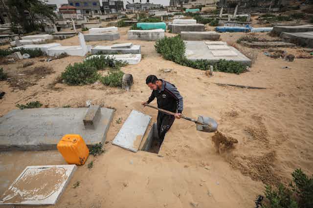 A man stands in a hole, shoveling dirt out, surrounded by grave markers.