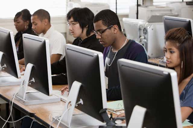 Five students of various ethnicities work at computers that sit atop a series of tables in a classroom.