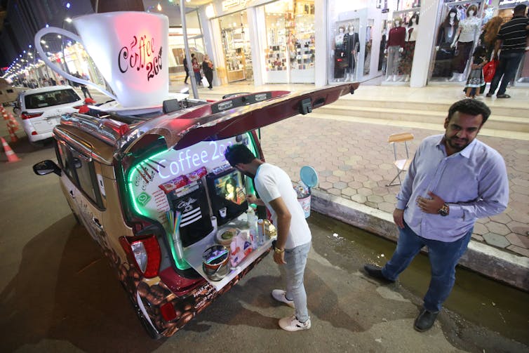 A man makes a coffee at a machine in the back of a small vehicle, while another man waits.