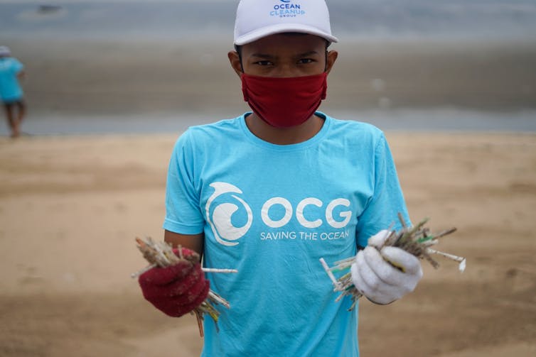 A person stands holding plastic straws at the beach
