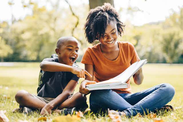 A smiling boy points to a page in a book being held by his older sister as they sit cross-legged in the grass in an outdoor space.