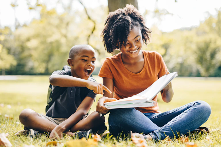 A smiling boy points to a page in a book being held by his older sister as they sit cross-legged in the grass in an outdoor space.