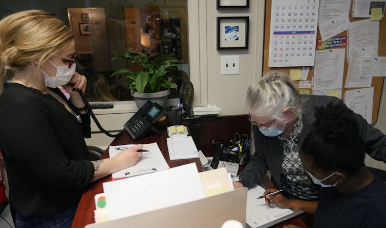 Two women wearing masks stand over a crowded office desk with papers and answer a landline phone.