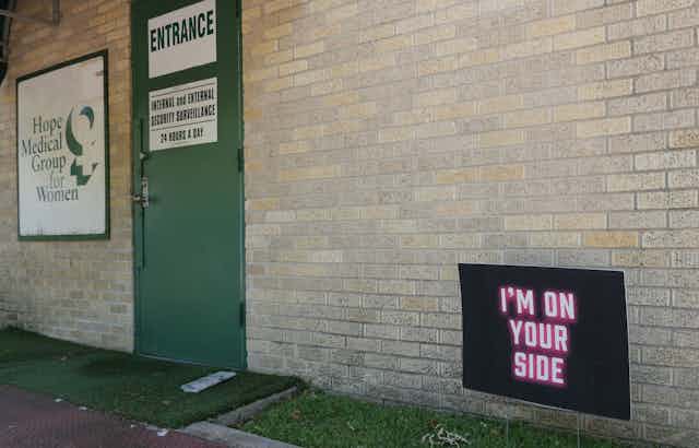 A neon sign in pick says 'I am on your side' in front of a brick building with a green door that says entrance and a sign for the Hope Medical Group for Women