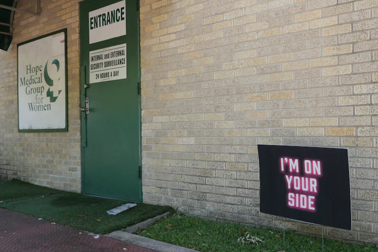 A neon sign in pick says 'I am on your side' in front of a brick building with a green door that says entrance and a sign for the Hope Medical Group for Women