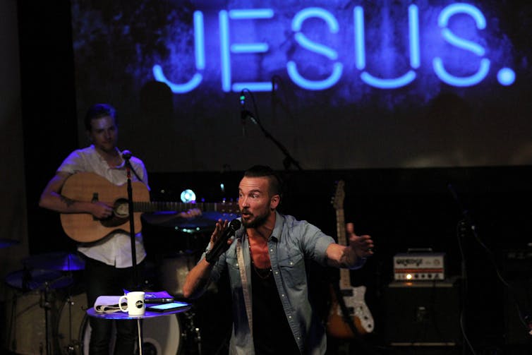 A bearded man in an open shirt speaks into a microphone under a neon light reading 'JESUS'
