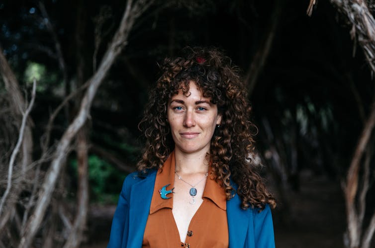 A woman with long, curly hair and a slight smile, standing against trees