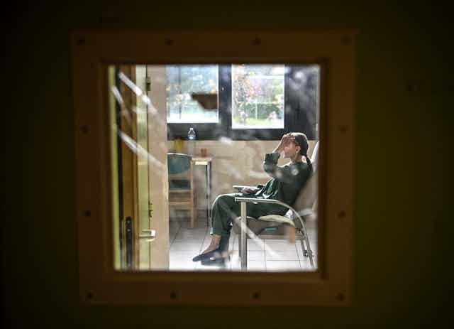 Une patiente est assise sur chaise dans sa chambre et on la voit derrière la vitre de sa porte fermée.