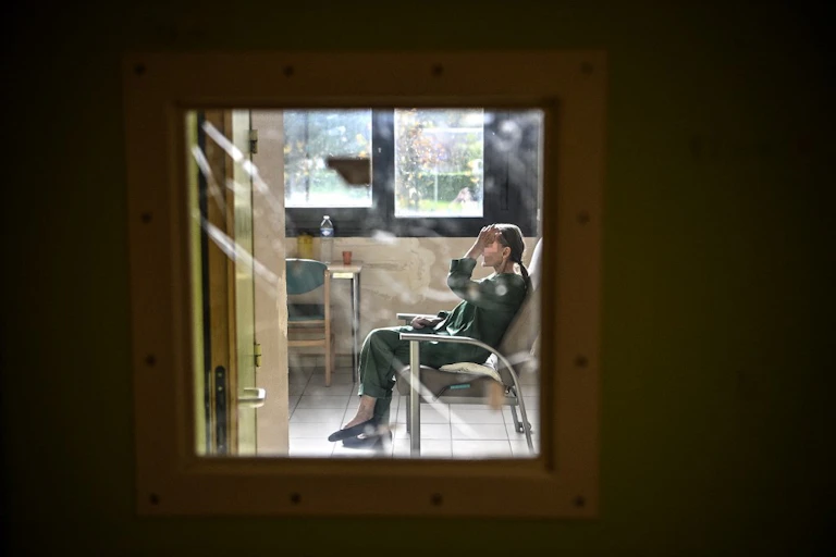 Une patiente est assise sur chaise dans sa chambre et on la voit derrière la vitre de sa porte fermée.