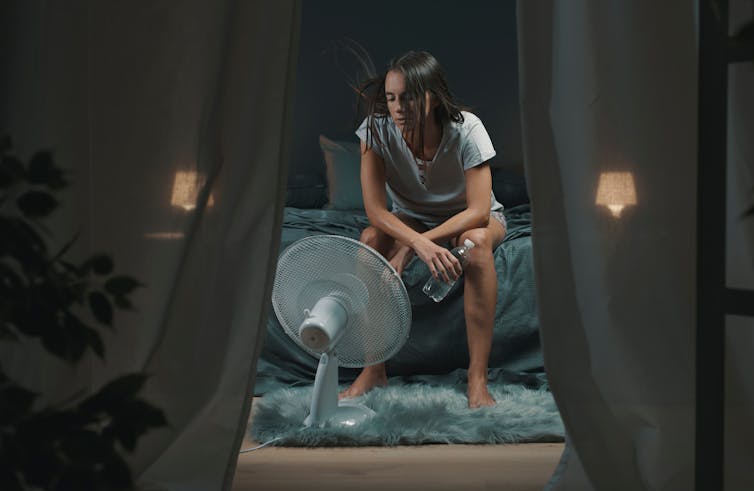 A woman sits on a bed in front of an electrical fan.