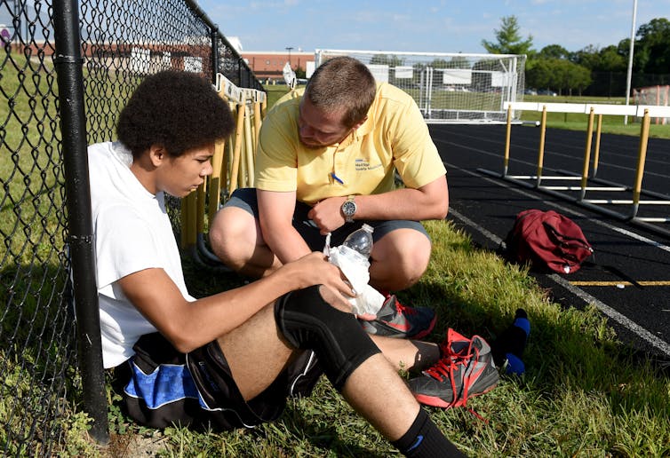 A young player sits against a fence next to a track looking exhausted while a man crouches down next to him and talks to him.