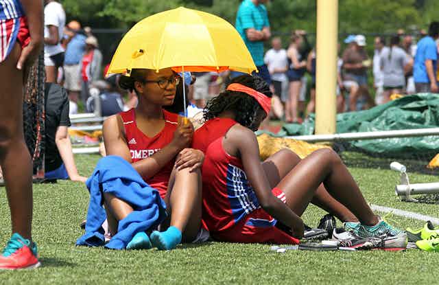 Young Black female runners sit under a small hand-held umbrella with teammates in the background