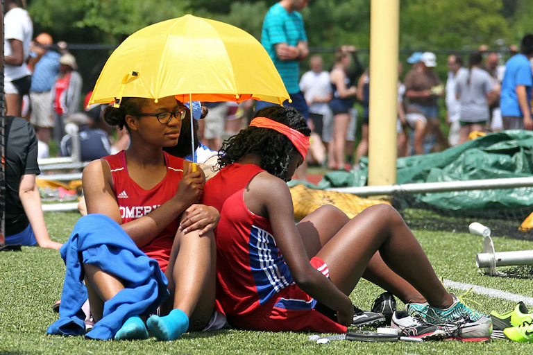 Young Black female runners sit under a small hand-held umbrella with teammates in the background