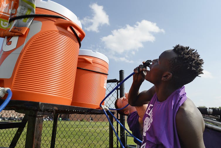 Two teenage players drink from large coolers near a playing field