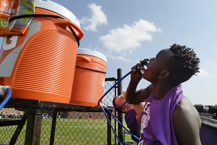 Two teenage players drink from large coolers near a playing field
