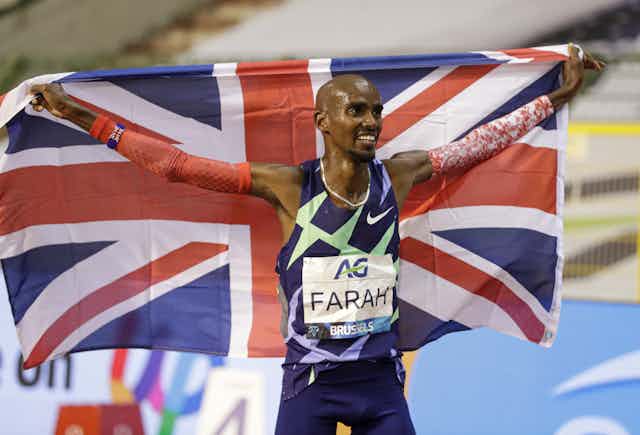 Mo Farah after a race, arms outstretched holding a UK flag behind him and smiling