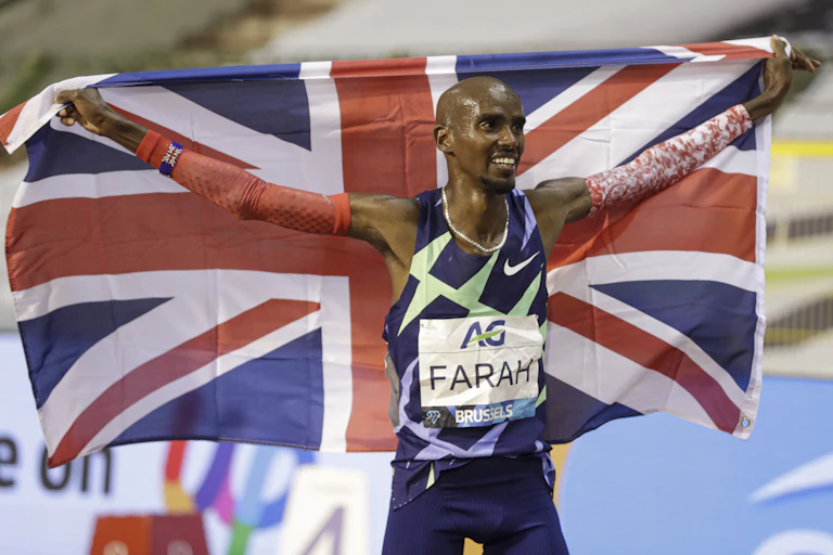 Mo Farah after a race, arms outstretched holding a UK flag behind him and smiling