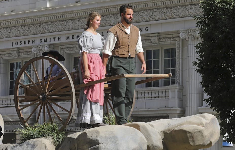 A young woman and man with a beard in old-fashioned pioneer clothing pose with a wooden handcart.